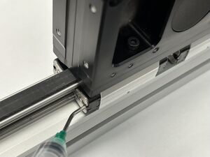 Insert angled syringe tip into grease hole in bearing end cap. Move the carriage about 50 mm while squeezing approximately 0.2 mL of grease from the syringe. Repeat for all eight end caps. Cycle the carriage through the full range of travel.